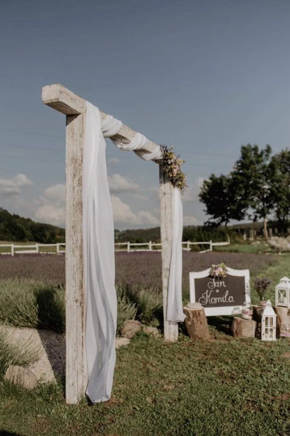 Wooden wedding arch with fabric draping, “Jan & Kamila” chalkboard sign, lanterns and lavender field