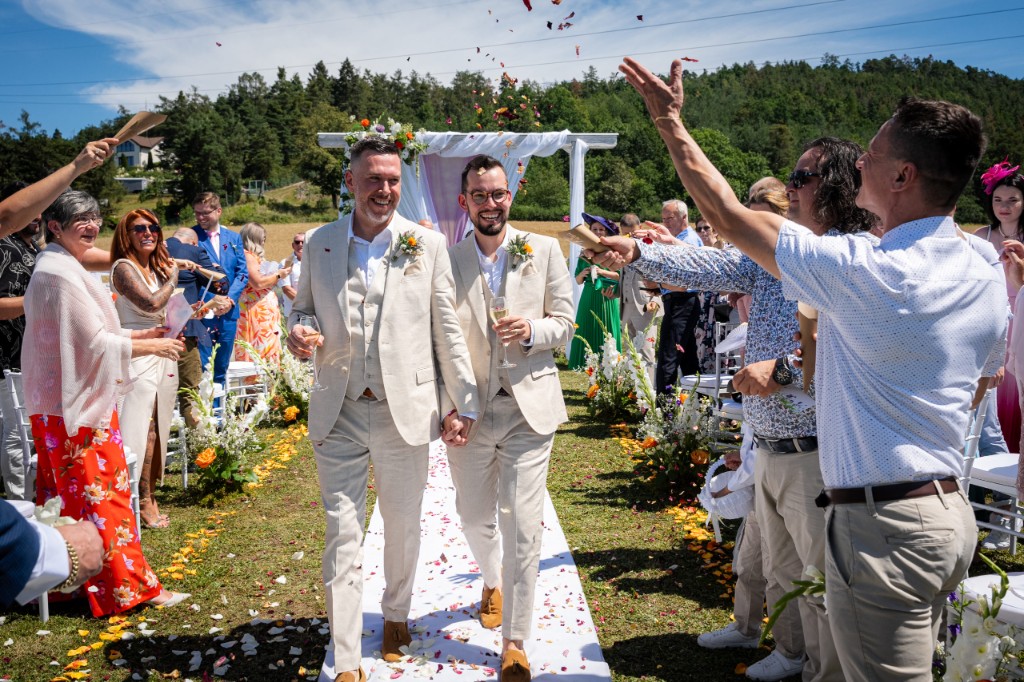 Guests throw petals as the grooms walk down the aisle with champagne